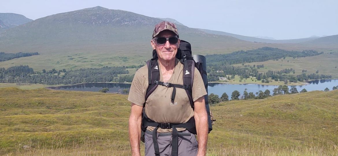 Man with rucksack on back with hills and loch behind.