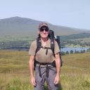 Man with rucksack on back with hills and loch behind.